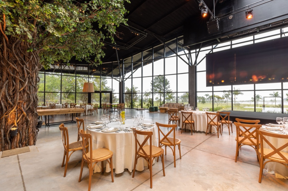 A glass-walled event hall set with round tables, wooden chairs, and a stage, in daylight.