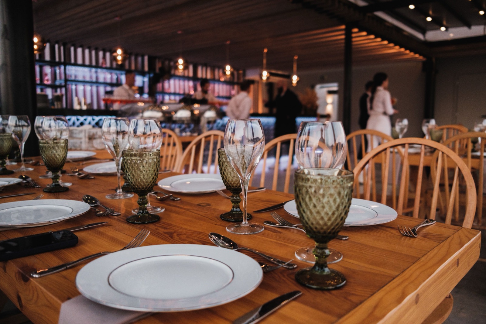 A set table in sharp focus with wooden chairs, while staff work the service line in the background.