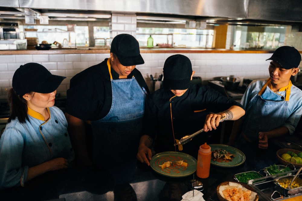 Four kitchen staff gather at the pass while one of them plates a dish.
