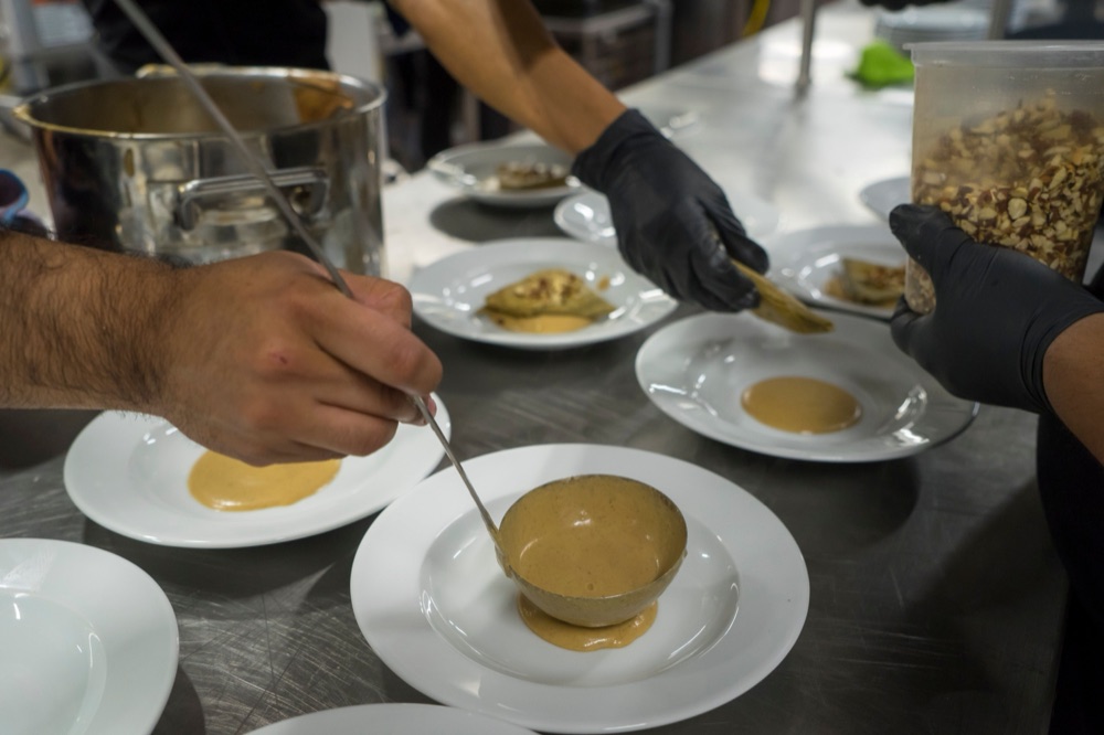 Three cooks plate dishes side by side on a steel pass, one ladling sauce.